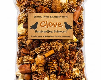 May include: A clear plastic bag filled with dried botanical potpourri, including pinecones, cinnamon sticks, and seed pods. A tan label reads: Shoots, Roots & Leather Boots, Clove, Handcrafted Potpourri, Proudly Made In Robertson County, Tennessee.