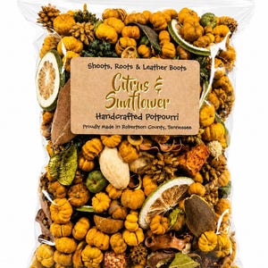 May include: A clear plastic bag filled with dried potpourri containing small orange pumpkins, green citrus slices, and brown botanical elements. A tan label reads: Shoots, Roots & Leather Boots, Citrus & Sunflower, Handcrafted Potpourri, Proudly Made in Robertson County, Tennessee.