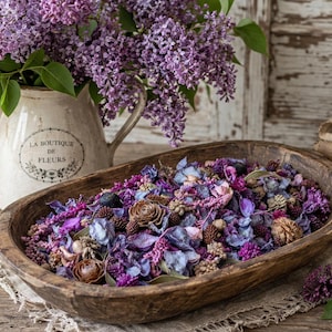 May include: A rustic wooden bowl filled with dried flowers and pinecones in shades of purple, blue, and brown. A cream-colored pitcher with purple lilacs and the text "LA BOUTIQUE DE FLEURS" is in the background. The arrangement sits on a wooden surface.