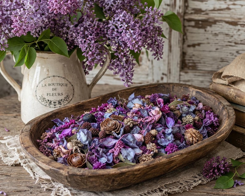 May include: A rustic wooden bowl filled with dried flowers and pinecones in shades of purple, blue, and brown. A cream-colored pitcher with purple lilacs and the text "LA BOUTIQUE DE FLEURS" is in the background. The arrangement sits on a wooden surface.