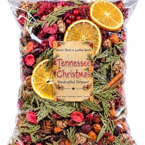 May include: A clear plastic bag filled with potpourri containing dried orange slices, green cedar sprigs, pinecones, red berries, and cinnamon sticks. A label reads: Shoots, Roots & Leather Boots, Tennessee Christmas, Handcrafted Potpourri, Proudly Made in Robertson County, Tennessee.