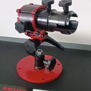 May include: A red metal tool stand with a black metal tool attached. The tool has a black handle and several black knobs. The stand has a round base with the words "ROUTE TOOL PRO" printed on it.