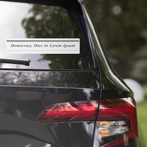 Puede incluir: Una pegatina rectangular blanca con el texto "Democracy Dies in Lorem Ipsum" en negro está adherida a la ventana trasera de un coche gris oscuro. El piloto trasero del coche es visible.