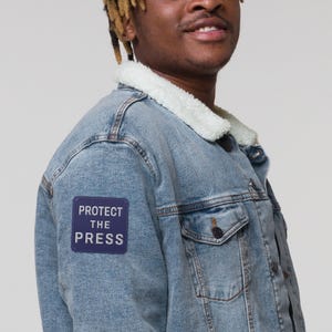 May include: Light blue denim jacket with a white faux shearling collar. A navy blue patch on the left breast pocket reads "PROTECT THE PRESS" in white capital letters. The jacket has two chest pockets and a button closure.