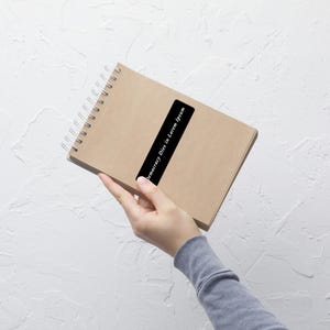 Puede incluir: Un cuaderno con espiral y cubierta de papel kraft. Una pegatina rectangular negra con el texto "Democracy Dies in Lorem Ipsum" está adherida diagonalmente a la cubierta. El cuaderno se sostiene sobre un fondo blanco texturizado.