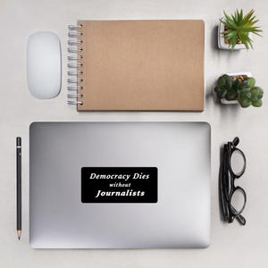 Puede incluir: Una pegatina para portátil con el texto "Democracy Dies without Journalists" en blanco sobre un rectángulo negro. La pegatina está sobre un portátil plateado, junto a un ratón blanco, un cuaderno marrón, un bolígrafo, gafas y pequeñas plantas en macetas.