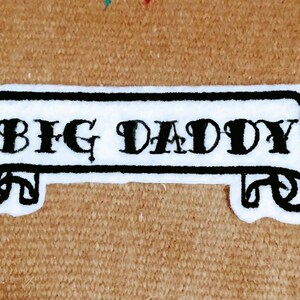 Puede incluir: Parche blanco bordado con puntadas negras. El parche presenta una pancarta con el texto "BIG DADDY" en una fuente estilizada.