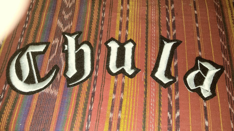 May include: A close-up of a fabric patch with the word "Chula" written in white letters on a black background. The letters are in a gothic font style.