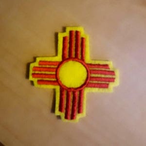 May include: A yellow and red embroidered patch featuring the New Mexico Zia symbol. The patch has a yellow border, a yellow circle in the center, and red lines and shapes. The Zia symbol is a Native American symbol representing the sun.