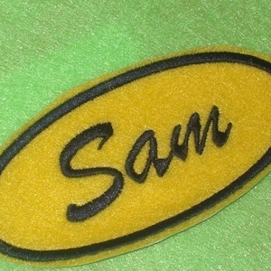 May include: A yellow oval-shaped patch with a black outline and the name "Sam" written in a black cursive font.