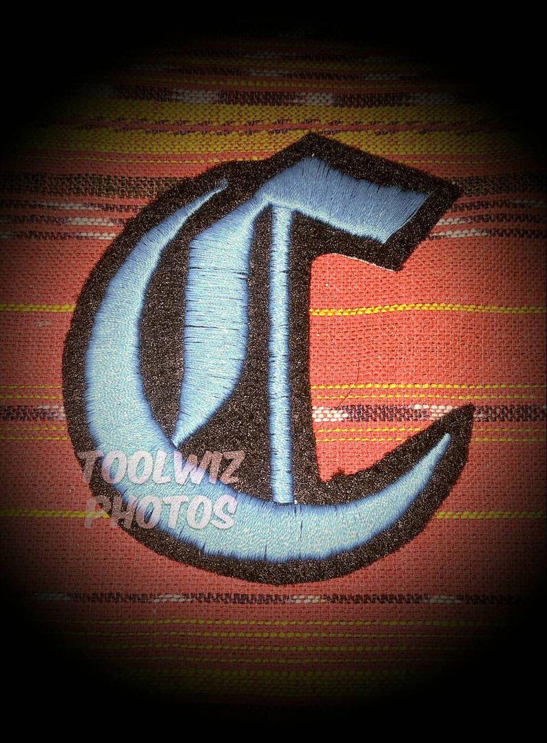May include: A blue and black embroidered letter C patch. The patch is on a red and yellow striped fabric background.