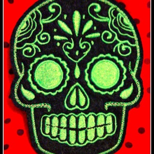 May include: A black and green embroidered sugar skull patch. The skull has a detailed design with swirls and flowers.