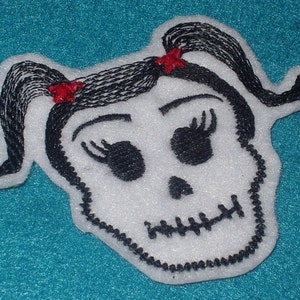 Puede incluir: Calavera bordada blanca con pelo negro y lazos rojos, la calavera tiene pestañas y una boca cosida.