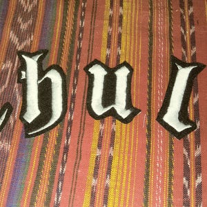 May include: White felt letters spelling out the word "Chula" in a gothic font, sewn onto a striped fabric background.