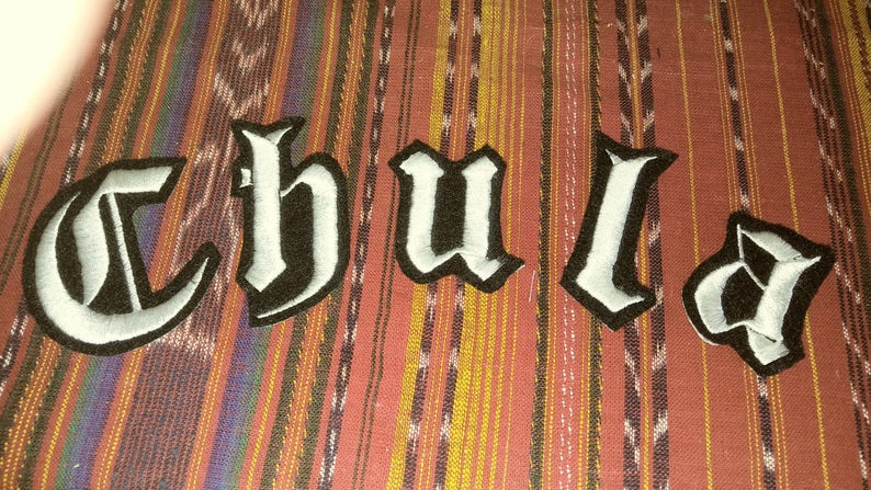 May include: White felt letters spelling out the word "Chula" in a gothic font, sewn onto a striped fabric background.