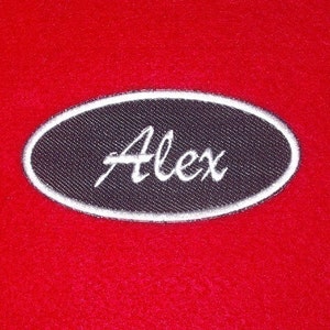 May include: A white oval patch with a black background and the name "Alex" written in white cursive lettering.