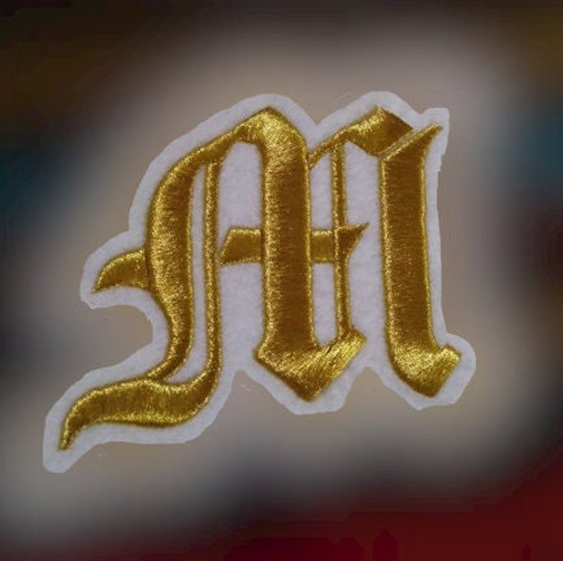 May include: Gold embroidered iron-on patch of the letter "M" in a gothic font style. The patch is on a white background.