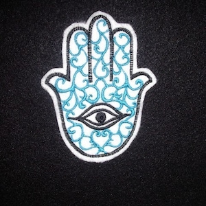 Könnte beinhalten: Eine gestickte Hamsa-Hand in Blau und Weiß mit einem Auge in der Mitte. Die Hamsa-Hand ist ein Symbol für Schutz und Glück.