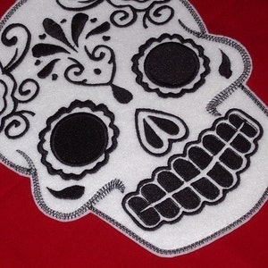 May include: White sugar skull with black embroidery details. The skull has a black eye socket, a black heart shape, and a black mouth with teeth.