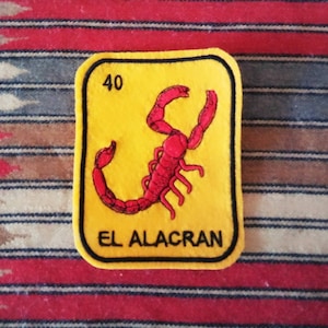 May include: A rectangular yellow patch with a black border featuring a red scorpion illustration in the center. The number 40 is printed in the top left corner, and the text EL ALACRAN is printed in black capital letters at the bottom.