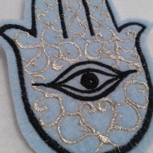 May include: A light blue felt patch with a black outline of a hamsa hand with a silver eye in the centre. The hamsa hand is a symbol of protection and good luck.