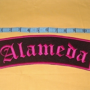 Peut inclure: Un écusson brodé noir et rose avec le mot "Alameda" en police gothique.