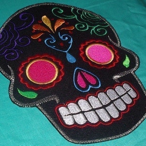 May include: Black embroidered sugar skull with colorful details, including pink, green, blue, and yellow. The skull has a white smile and is surrounded by a silver trim.