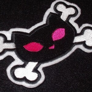 May include: Black and white embroidered patch of a cat skull with pink eyes. The cat skull is surrounded by white bones.