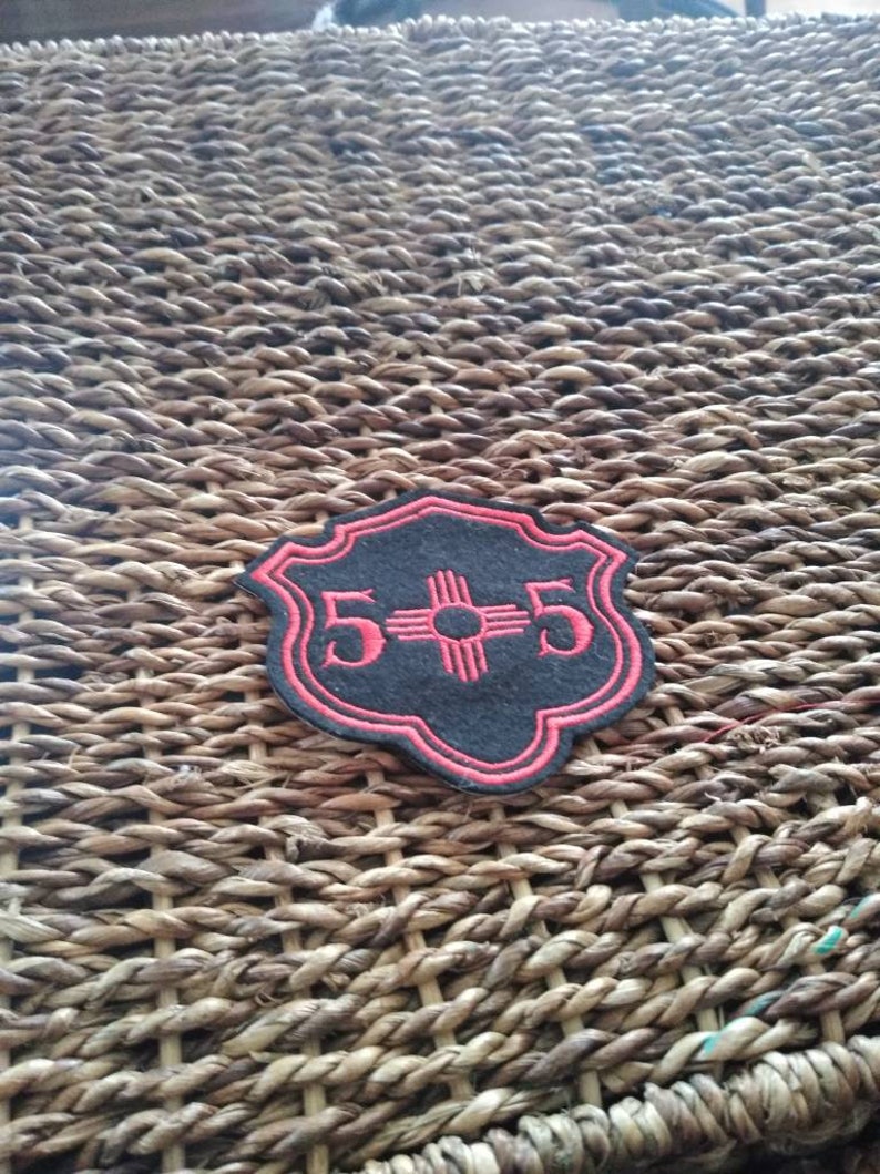 May include: A black and red patch with the text "5+5" and a stylized sun symbol. The patch has a decorative border and is set against a woven background.
