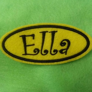 Könnte beinhalten: Gelber Filz-Oval mit schwarzem Rand und dem Namen "Ella" in schwarzer Stickerei.