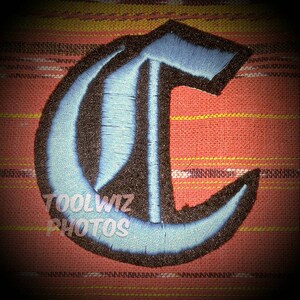 May include: An embroidered letter C in a gothic font style, with blue and black stitching.