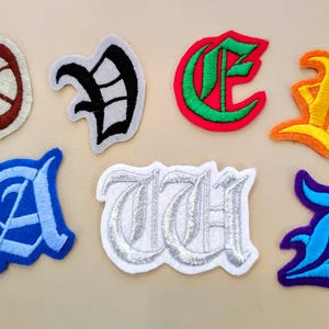 May include: A collection of seven embroidered patches featuring various letters and symbols in gothic-style typography. The patches include a brown and white circular design, a black letter, a green and red letter, an orange and yellow letter, a blue letter, a silver letter, and a light blue and purple letter.