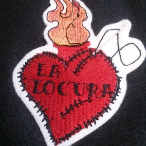 Peut inclure: Un cœur en feutrine rouge avec des coutures noires autour des bords. Le cœur est en feu et porte les mots "La Locura" brodés dessus.
