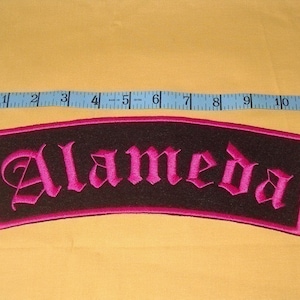 Peut inclure: Patch brodé rose avec le mot "Alameda" en noir sur fond noir. Le patch a une bordure rose et mesure environ 25 cm de large.