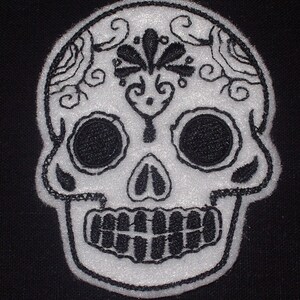 Puede incluir: Parche bordado de calavera de azúcar blanca con hilo negro. La calavera tiene diseños intrincados alrededor de los ojos y la frente.