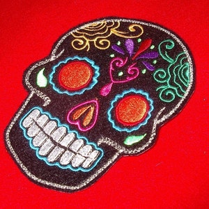 May include: Black embroidered sugar skull patch with colorful details, including red, orange, yellow, green, blue, and purple. The skull has a white smile and is surrounded by a silver outline.