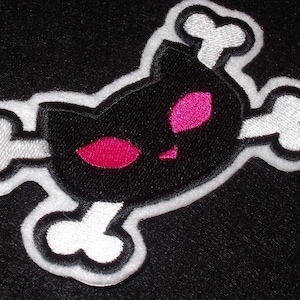 以下が含まれることがあります： 黒と白の刺繍パッチで、ドクロと交差した骨のデザイン。ドクロはピンクの目をした猫の形をしています。