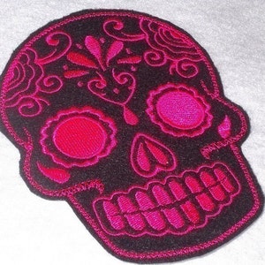 May include: Black felt sugar skull patch with pink embroidered details. The skull has a floral design on the forehead and pink eyes, nose, and mouth.