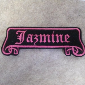 Może przedstawiać: Czarna i różowa haftowana naszywka z napisem "Jazmine" w formie zwiniętego zwoju.