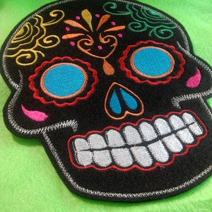 May include: Black felt embroidered sugar skull patch with colourful details, including swirls, flowers, and eyes. The skull is smiling with white teeth.