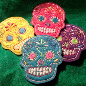 Puede incluir: Cuatro parches de calavera de azúcar de fieltro bordados en diferentes colores: amarillo, rosa, azul verdoso y morado. Cada calavera tiene detalles bordados, incluidos ojos, nariz y boca.