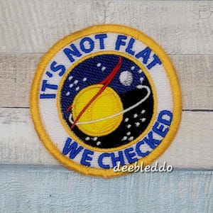 May include: A circular embroidered patch with a yellow border. The center features a yellow planet, a red trajectory line, and a white ring against a dark blue starry background. The white border contains the blue text IT'S NOT FLAT WE CHECKED.