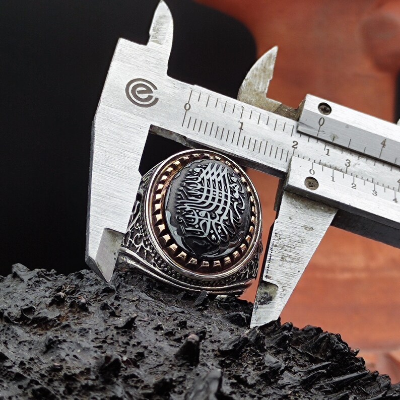 May include: A silver ring with a black onyx stone and intricate detailing. The stone features Arabic calligraphy in gold. The ring is being measured with calipers.