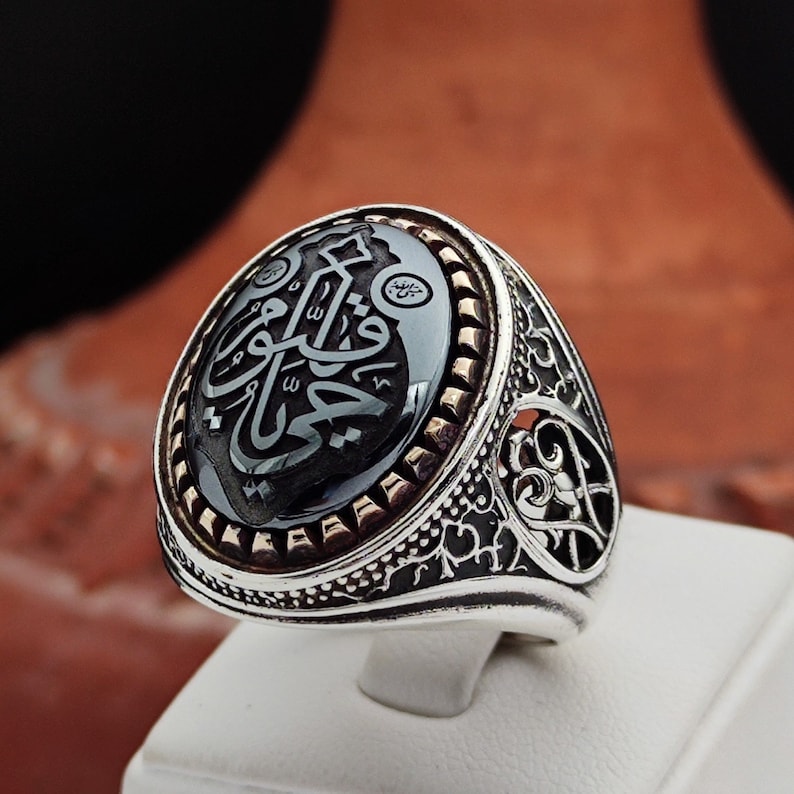 May include: A silver ring with an intricate design. The ring features a black onyx stone with Arabic calligraphy. The stone is surrounded by a gold-colored band.