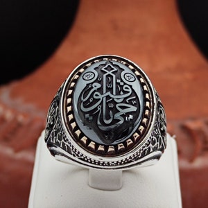 May include: A silver ring with an ornate design. The ring features a black onyx stone with Arabic calligraphy in gold. The stone is surrounded by a silver band with a decorative border.