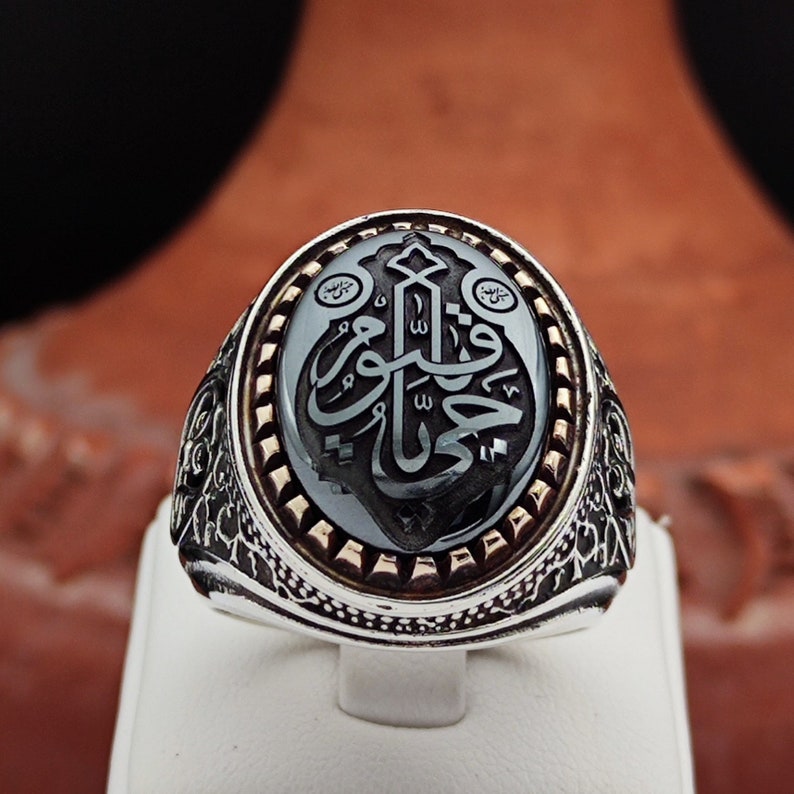May include: A silver ring with an ornate design. The ring features a black onyx stone with Arabic calligraphy in gold. The stone is surrounded by a silver band with a decorative border.