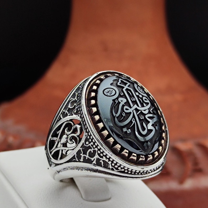 May include: A silver ring with an oval black onyx stone featuring Arabic calligraphy. The ring has a detailed filigree design and a gold-toned border around the stone.