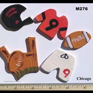 May include: A set of five ceramic painted football-themed ornaments. The ornaments include a football helmet, a football jersey, a football, a pair of football pants, and a football in a goal post. The ornaments are painted in red, white, black, brown, and green. The ornaments are labeled "M276" and "Chicago".