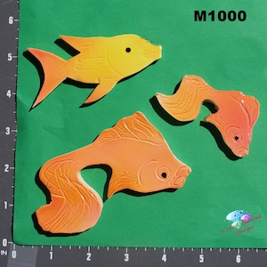 Puede incluir: Tres azulejos de cerámica en forma de pez, en tonos naranja y amarillo. Los azulejos tienen forma de pez dorado y una superficie texturizada. Los azulejos están etiquetados como "M1000".