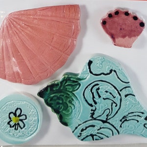 May include: Four ceramic tiles with a sea theme. One tile is a pink seashell, another is a blue seashell with black swirls, a third is a blue circle with a white flower, and the last is a small pink seashell with black dots.